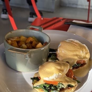 Two eggs benedict sandwiches with spinach and bacon on a plate, alongside a bowl of roasted potatoes