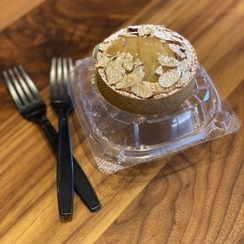 A round pastry with almond topping in a clear plastic container on a wooden surface, with two forks beside it