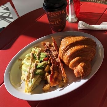 Breakfast plate with scrambled eggs, bacon, and croissant on red table with coffee cup