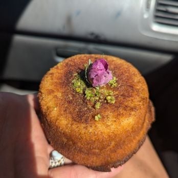 Golden-brown pastry held in hand, topped with pistachio crumbs and a pink crystallized flower