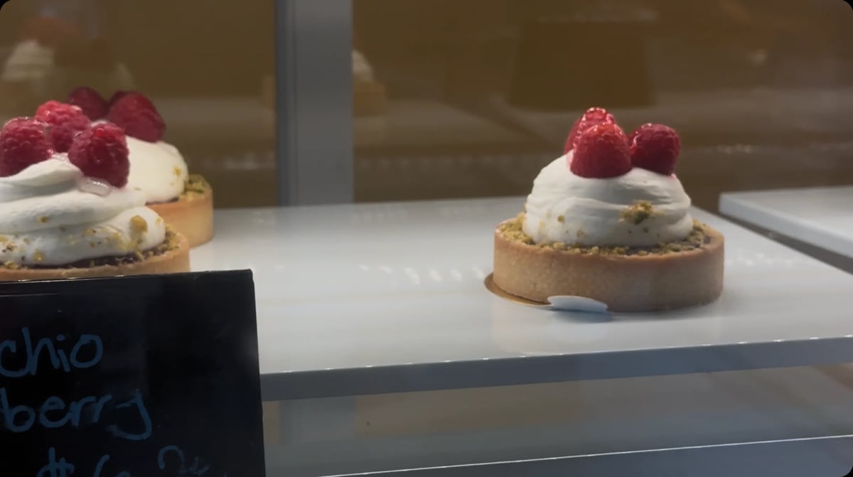 Two berry tarts with whipped cream and raspberries on display in a bakery case with a chalkboard menu label